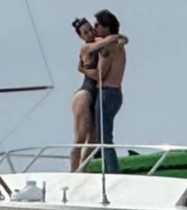 A man and woman stand embracing on the deck of a boat at sea, with the woman wearing a black swimsuit and her hair in a bun, wrapping her arms around the shirtless man who has long hair and is kissing her cheek, both positioned near a sail and railing with ocean waves and a green object visible in the background.